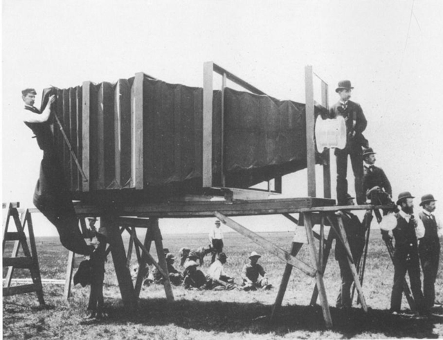 an old-tin camera with a bellows structure to distance the lens from the glass plate inside. It's on a purpose-built tower about 6 feet off the ground, and is taking a picture. The lens cover is as large as a trashcan lid.