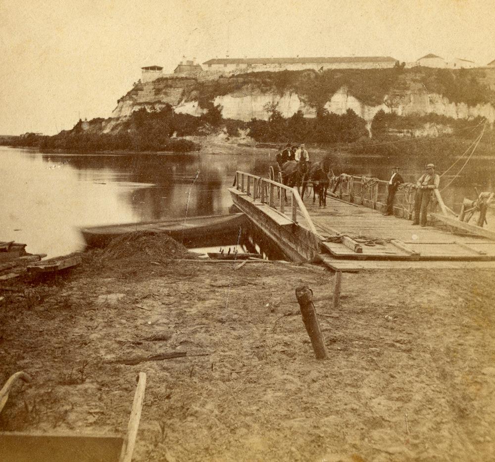 A fire of Fort Snelling in the distance with a flatbottomed ferry boat on the near shore. A wagon with a couple of people in it was on board, and a two ferrymen lounged against the ferry's railings.